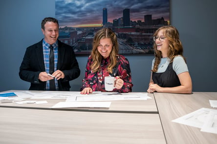 Three LX Studio team members collaborate around a conference table with papers and a coffee mug, smiling as they review project materials together.