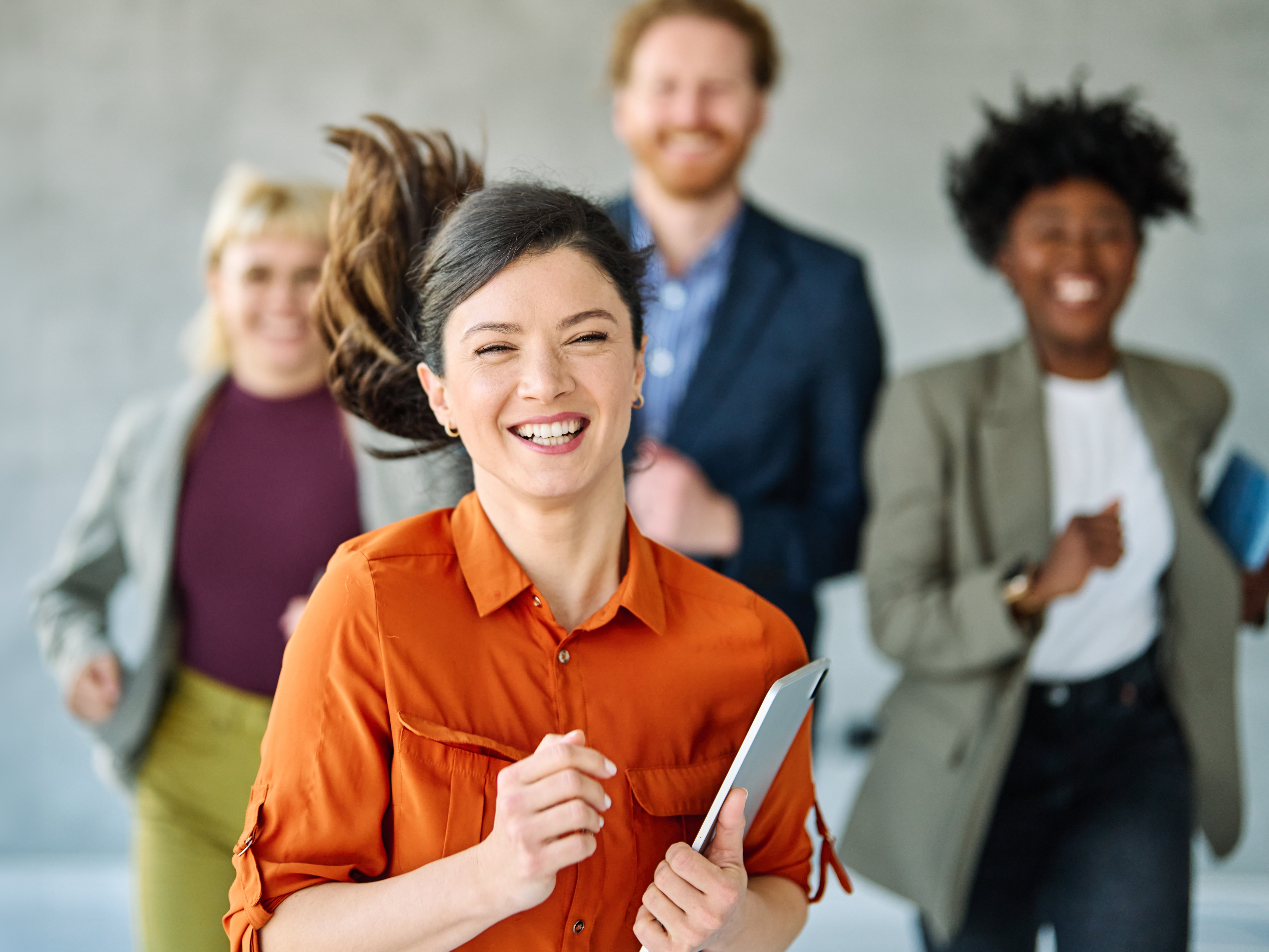 Smiling professional holding a tablet runs toward the camera with coworkers behind in an office.